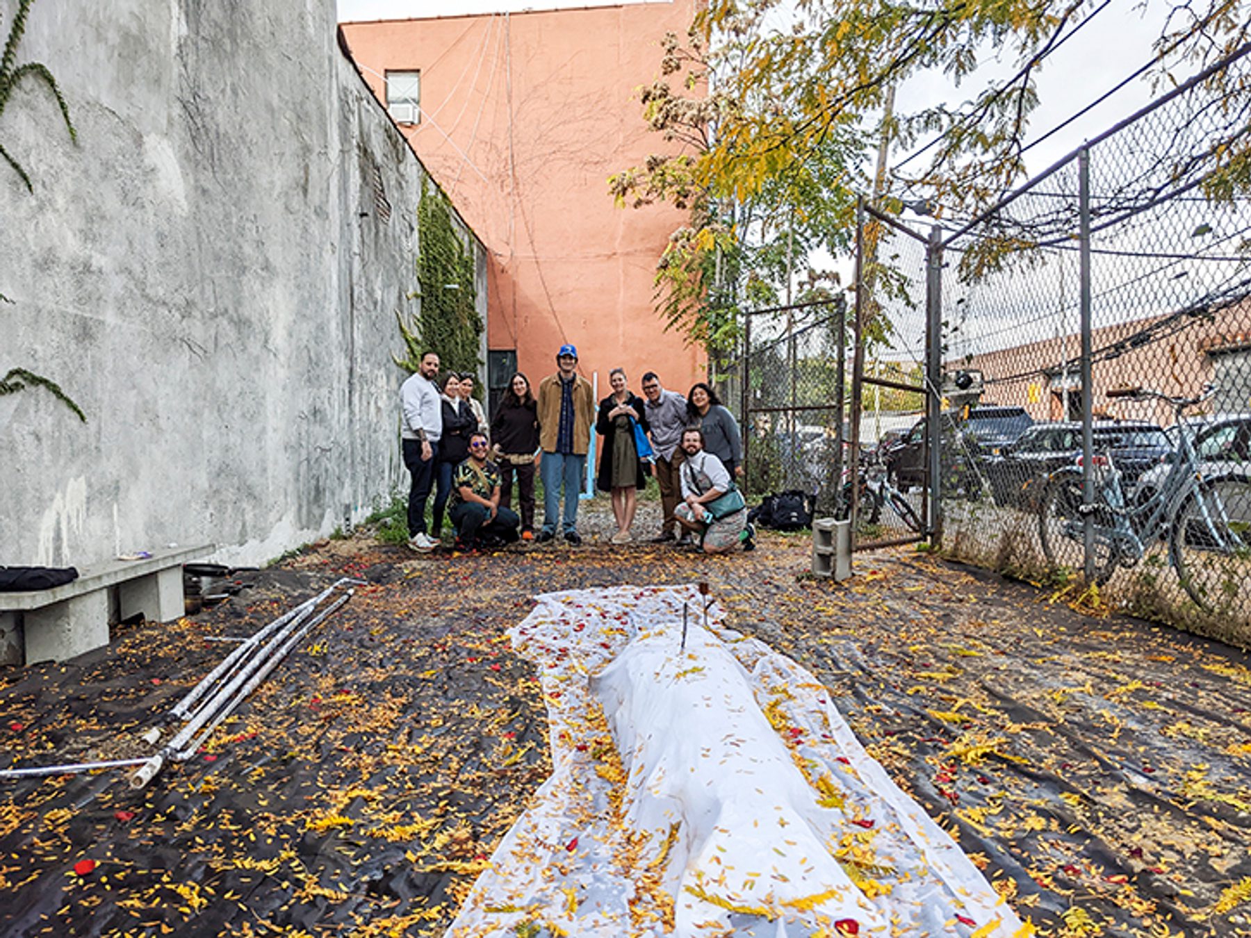 MFA students pose with Jacob Jackmauh and Lina McGinn in a fenced in area with fallen leaves and a gray, concrete wall.
