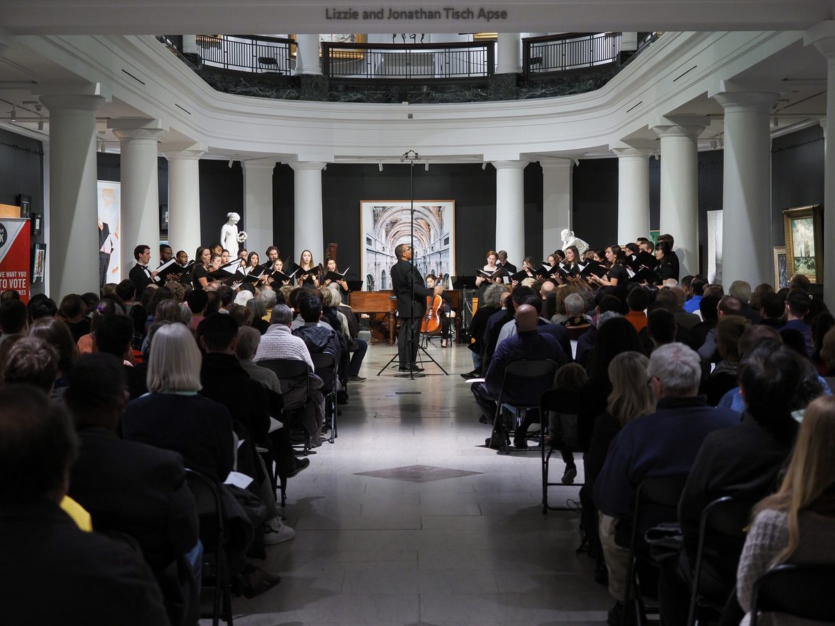 Image of a performer standing in a large room in a museum as an audience looks on.