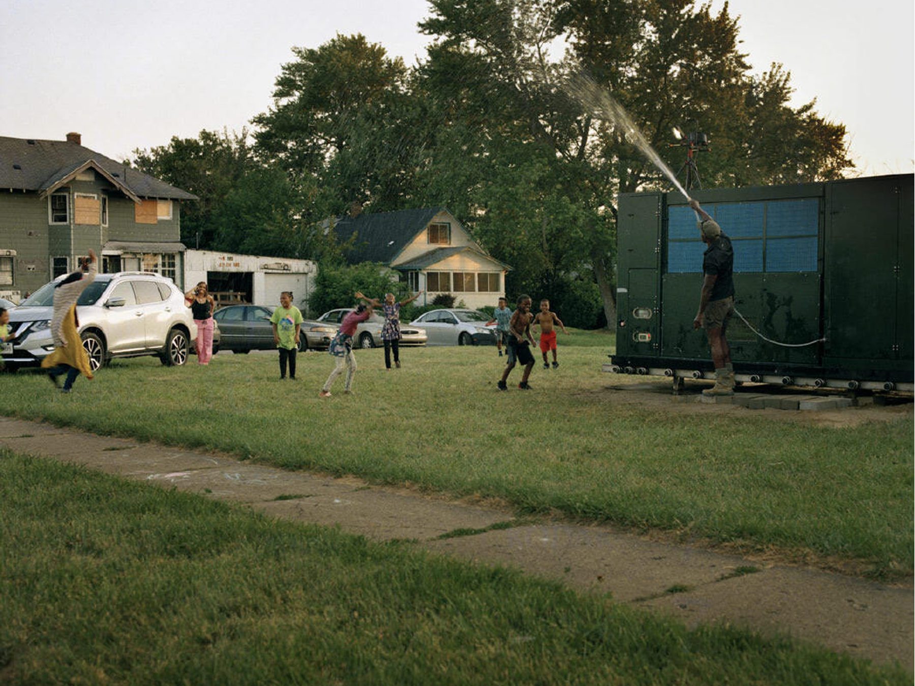 Photograph of a group of people in a grassy lot playing in water that is being sprayed from a large truck with solar panels