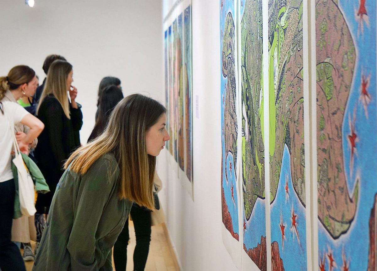 Viewers inspect Endi Poskovic's colorful large scale prints on view in a white-walled, bright gallery