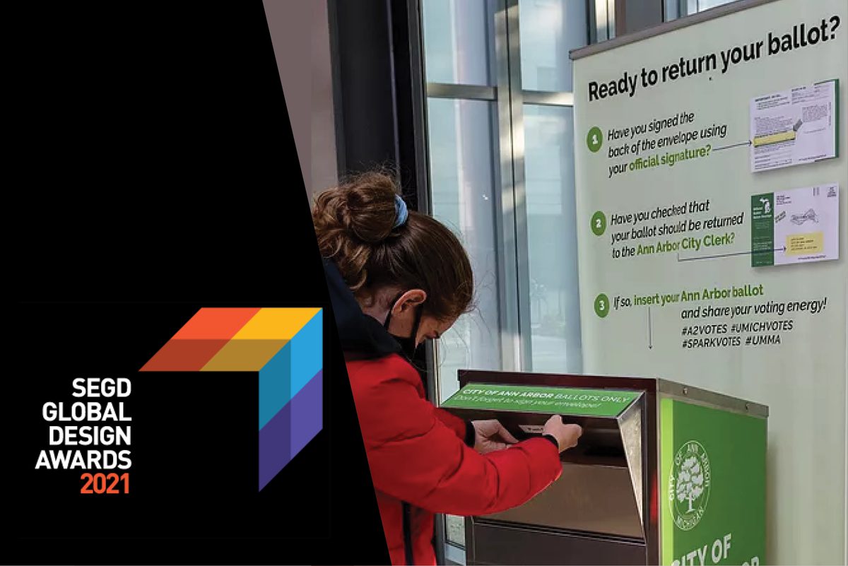 A pony tailed student drops an absentee ballot into a mail box. This image also contains the SEGD Global Design Awards logo.