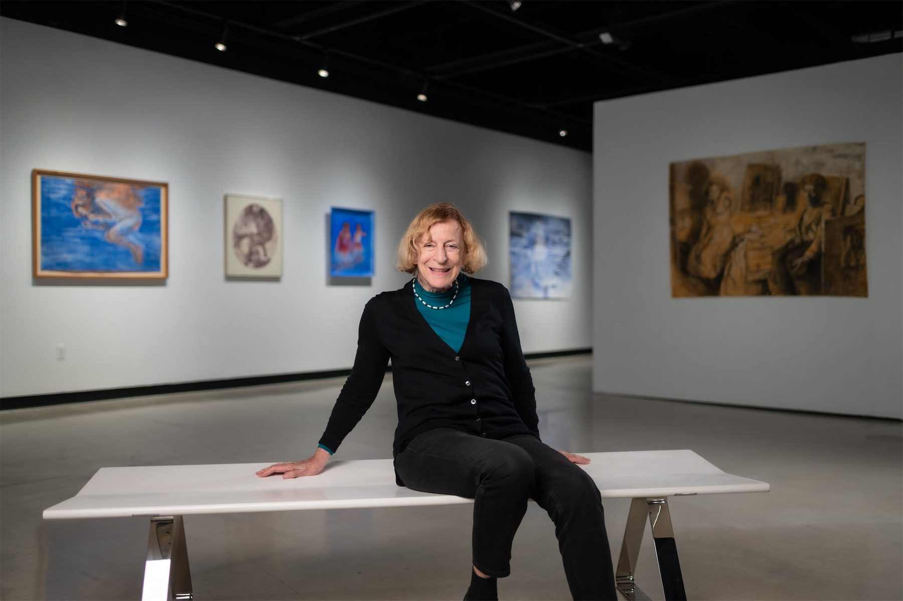 Ruth Weisberg, wearing a black sweater and green shirt, sits on a white bench in Stamps Gallery, surrounded by her artwork.