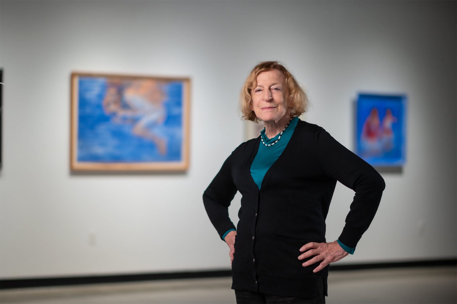 Ruth Weisberg, wearing a black sweater and green shirt, stands, hands on hips, surrounded by her artwork.