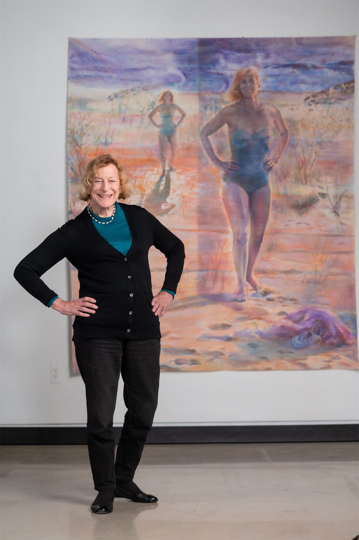 Ruth Weisberg, wearing a black sweater and green shirt, poses with hands on hips in reference to a painted self portrait on the wall