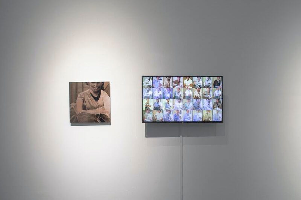 On a white gallery wall, a large print of a strangely disfigured human is shown next to a monitor with dozens of faces displayed on it