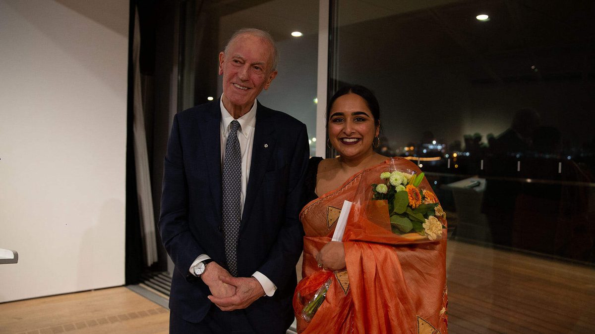 Simranpreet Anand, wearing a bright orange sari and holding a bouquet of flowers, stands with Philip B. Lind
